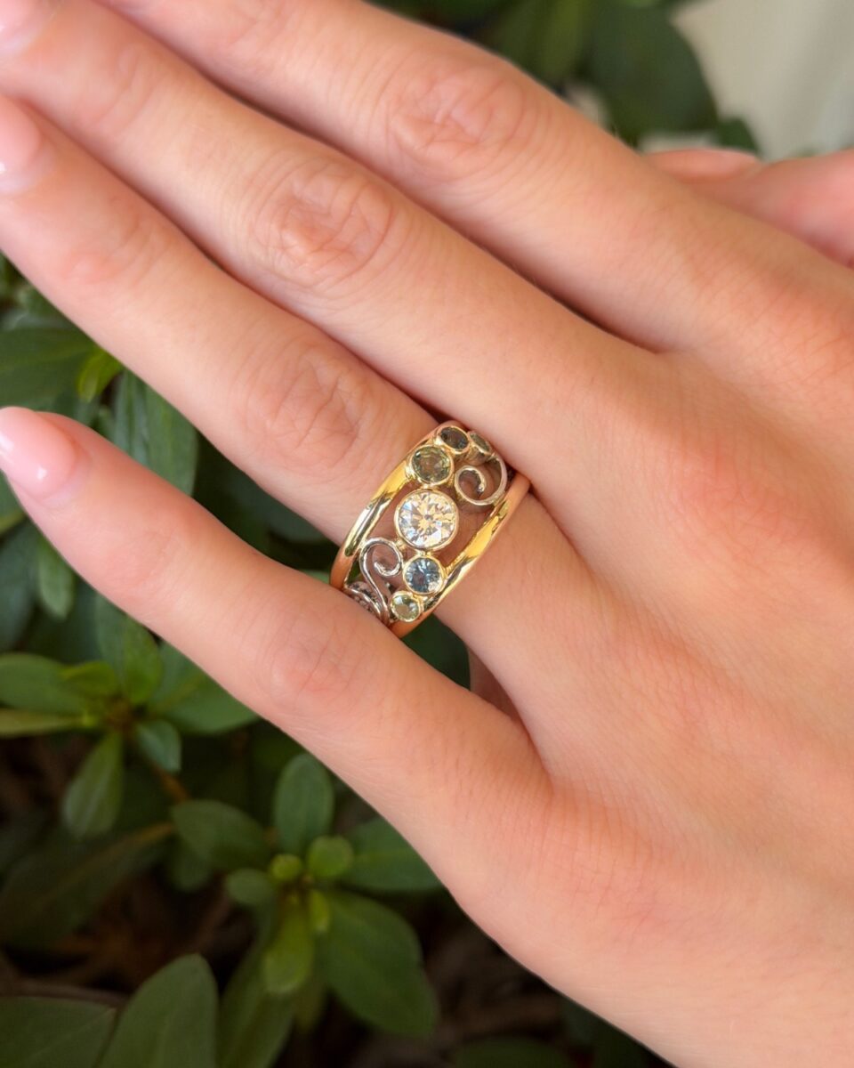 Finished gem-set band worn on the hand, with a centre diamond, green and blue sapphires, peridot and white gold scrollwork