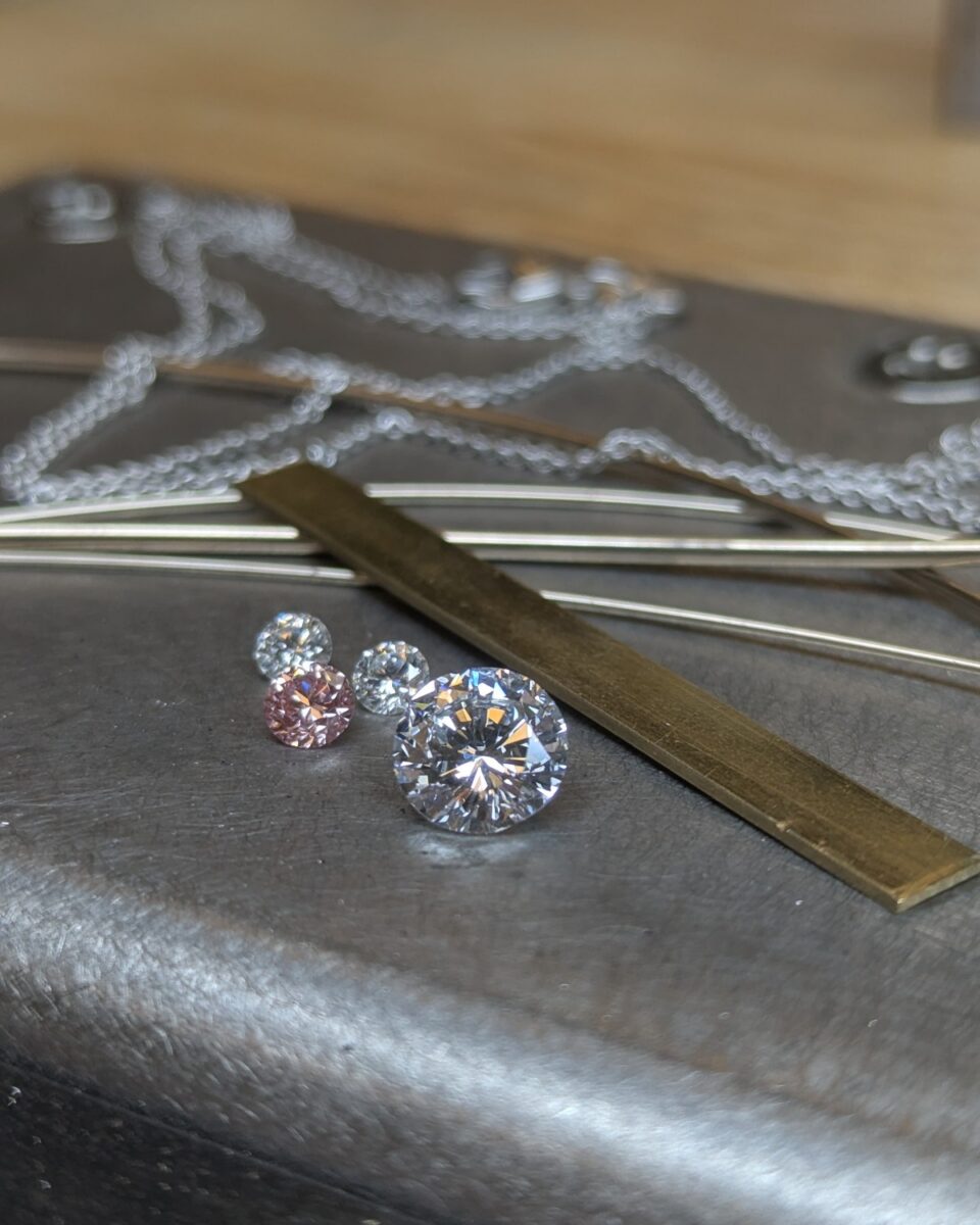 Loose diamonds and a pink sapphire beside gold wire and chain on the bench block, before fabrication begins